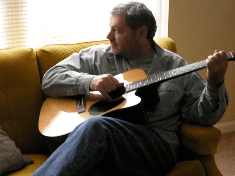 A man sitting on the couch holding an acoustic guitar.